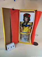 Poly commemorative doll in original yellow and red box with protective foam inside, doll dressed in yellow with blue and red trim, red headband.