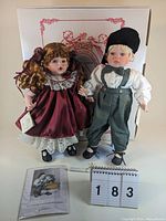 Two Fayzah Spanos heirloom dolls named Kisses & Jacques Love at First Sight, circa 1987, measuring 15 inches tall, standing in front of a box with floral border.