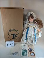 Photo shows the 20 inch Robin Woods doll named Roberta dressed in white lace and ribbons, her face and outfit clearly visible next to original cardboard box with Robin Woods logo and two pamphlets, also displaying small accessories or doll parts in plastic bag.