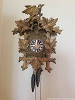Front view of whole cuckoo clock with bird on top and leaf carvings around clock face, showing pinecone weights hanging.