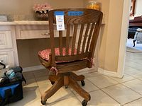 Rear angle view of vintage oak desk chair showing tall slatted backrest, cushioned seat, swivel base, and caster wheels. Blue tape with measurements visible.
