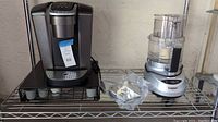 Keurig coffee maker with black pod storage drawer and Cuisinart Prep 7 food processor with clear plastic bowl and accessories on a metal shelving rack.