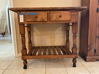 Front view of solid wood kitchen cart showing two drawers, turned legs, slatted shelf, and caster wheels.