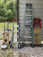 Overview of all five items leaning against metal wall: three ladders, hand truck and platform dolly