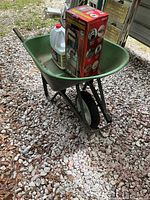 Green metal wheelbarrow with a box labeled Gilmour Professional sprayer and a container of outdoor cleaner inside it, positioned outdoors on gravel.