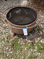 Full view of round metal outdoor fire pit with black grill grate, standing on three legs on gravel surface.