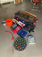 Photo shows overall lot items scattered on garage concrete including stadium seats, blankets, bags, orange bucket, metal pot, dartboard, and others.