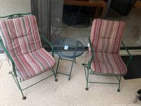 Two green metal framed outdoor chairs with red and beige striped cushions and one round blue metal mesh side table arranged on a patio floor.