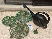 Photo showing three green cast iron planter stands of different sizes and a black plastic watering can on a concrete patio.