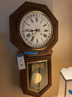 Front view of wooden regulator wall clock with visible face showing Roman numerals and day of month markings, glass front on pendulum box with 'REGULATOR' text.
