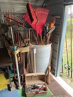 Photo showing yard tools including large red plastic rake, metal rakes, shovels, and a broom stored in and around a white plastic barrel and beige tool holder stand.