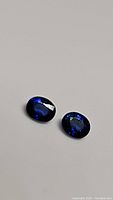 Two small oval royal blue Ceylon sapphires placed side by side on a neutral background showing color and shape.