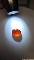Oval cabochon carnelian gemstone illuminated by flashlight on a white surface, showing glossy smooth polished surface and bright orange color.