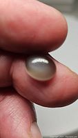Close-up of the 3.67 carat natural oval cabochon grey silver moonstone held between fingers showing its translucence and adularescence.