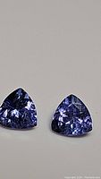 Two close-up images of a pair of trillion cut deep blue-purple tanzanite gemstones laid flat on a neutral surface, highlighting their color and faceting.