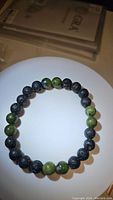 Photo of the bracelet laid flat showing alternating green jade and black lava stone beads arranged in a circular pattern on a white surface.