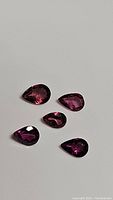Five pear cut natural pink garnet gemstones of varying size on a plain white background, showing natural faceting and bright pink color.