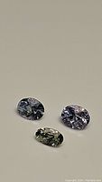 Three oval cut natural tanzanite gemstones laid on light surface; two appear purple-blue, one appears blue-green-yellow.