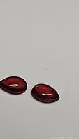 Pair of pear-shaped polished red-orange garnet cabochon gemstones placed on white background, showing color and smooth surface.