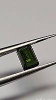 Photo of deep green rectangular emerald-cut tourmaline gemstone held in tweezers showing color and cut.