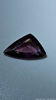 Photo of a deep purple, fancy trillion cut sapphire gemstone on a dark surface, showing its facets and shape.