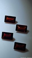 Four orange-red fancy baguette cut almandine garnet gemstones arranged on a flat surface under lighting that reveals their cut facets and transparency.