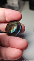 Close-up photo of a round labradorite gemstone with checkerboard cut faceting showing multicolored flash, held between fingers for scale.