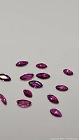 Close-up of multiple small marquis-cut natural rubies in shades of fuchsia, purple, pink and reddish tones, laid on white surface showing quality and size variations.