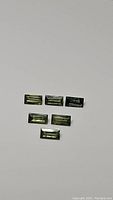 Six natural bright green baguette cut sapphires arrayed on a white surface, showing rectangular facets.