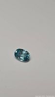 Close-up of 1.30ct oval cut lagoon blue zircon gemstone showing facets and transparency on a neutral background.