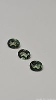 Three mint green oval cut apatite gemstones laid flat on a light surface, showing multiple facets and bright reflective qualities.
