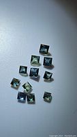 Overhead photo of 11 small princess cut multicolored sapphire gemstones showing blue, green, and teal tones on a light surface.