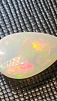 Close-up image of pear-shaped translucent white Ethiopian opal cabochon with multicolored flash on textured black background.