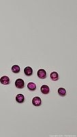 Close-up image showing thirteen round faceted rubies in various shades of exotic fuchsia colors ranging from purple to pink to red, arranged on a plain white background.