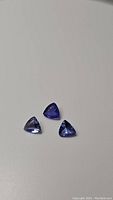Three natural trillion cut tanzanite gemstones in strong blue and purple colors on a white background.