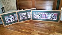 Three framed quilt storytelling prints displayed on floor in room with wood floors and chairs in background