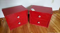 Front view of two red wooden chests with two drawers each and white ceramic apple knobs showing scratches and wear.