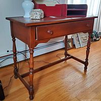 Antique maple wood desk showing the front and side, with single drawer and round knobs. Desk has turned legs and stretchers, stained finish.