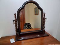 Front view of antique shaving mirror showing arched wooden frame, base with scratches and stains, and turned side supports with knobs for tilting.