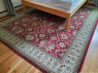 Full view of the oriental style rug under a wooden bed frame showing the scarlet red field and floral pattern with ivory floral border.