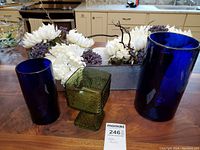 Shows two cobalt blue glass vases and one green glass compote with artificial white flowers in the background on a kitchen table.