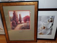 Both framed art prints leaning against a wall: scenic landscape and floral orchid photo