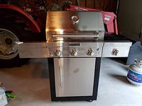 Front view of KitchenAid gas grill with four control knobs and side burner, stainless steel lid with temperature gauge, and propane tank to right side.