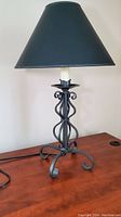Black table lamp with forged iron scrollwork base and black shade against neutral wall and wooden table.