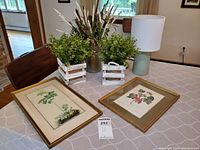 Photo of ceramic lamp with white drum shade, two framed botanical prints, two faux succulent plants in whitewashed wooden crates, and faux dried wild flowers in a green metal pitcher on a table