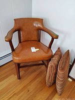 Front angle of brown leatherette armchair with curved wooden arms and nailhead trim, showing scratches on one arm. Two patterned decorative pillows next to the chair.
