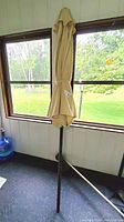 Canvas patio umbrella folded and secured with strap, standing indoors near window showing outdoor greenery.