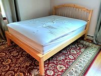 Full view of queen size pine wood bed frame with mattress on top, placed on an oriental rug near curtains.