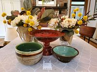 Wide photo showing all items on a table with pottery bowls, red footed fruit bowl, and three glass vases with floral arrangements.