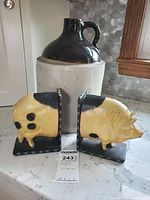 Pair of cast iron pig bookends on black L-shaped bases, positioned in front of a vintage stoneware whiskey jug, all placed on a kitchen counter with hexagonal tile backsplash.